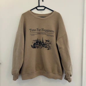 Beige sweatshirt med tryck - Beige sweatshirt med svart tryck framtill som visar en stadssilhuett och texten 'Time For Happiness'. Tröjan har rund hals, långa ärmar och ribbade muddar.
