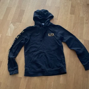 Svart EA7 hoodie med guldtryck - Svart hoodie från EA7 med snyggt guldtryck på bröstet och längs ena ärmen. Klassisk huva med snörning och stor magficka framtill. Perfekt för dig som gillar streetstyle och vill ha en stilren men ändå iögonfallande look.