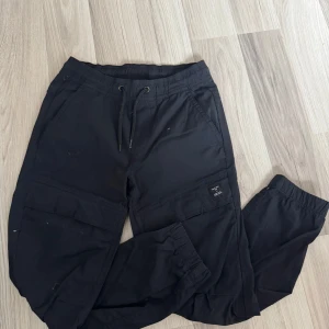 Svarta cargopants från lager 157 - Svarta cargopants från lager 157 med elastisk midja och snörning. Byxorna har flera fickor, inklusive stora benfickor, och är tillverkade i ett stretchigt material av 95% polyamid och 5% elastan. Perfekta för en avslappnad och cool stil.