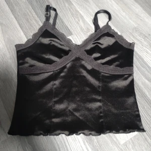 Sexigt, kort, Svart satin-linnetopp med spets H&M XS - Supersnygg svart linne-topp från H&M i satinliknande material med tunna axelband och spetsdetaljer vid bysten. Perfekt att styla med jeans eller kjol för en trendig look. Lätt glansig finish och vågig nederkant.