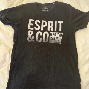 Svart t-shirt från Esprit med tryck - Svart t-shirt från Esprit med stort vitt tryck på bröstet där det står 'ESPRIT & CO REG.TM CALIF.'. Klassisk rund hals och korta ärmar. Perfekt för dig som gillar streetstyle och tydliga loggor.