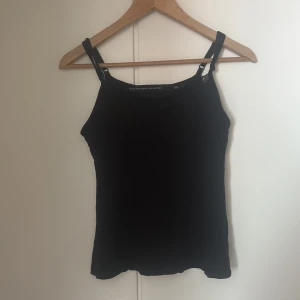 Svart linnetopp med spetsdetalj - Svart linnetopp från The Black Label Collection med smala axelband och snygga spetsdetaljer längs ringningen. Justerbara axelband för bästa passform. Strl M