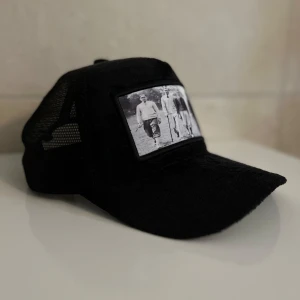 Svart keps med vintage fototryck - Svart keps med böjd skärm och mesh på sidorna. Framtill finns ett stort svartvitt fototryck som visar fyra personer i retrostil. Kepsen har klassisk truckerform och passar dig som gillar streetwear med en unik twist.