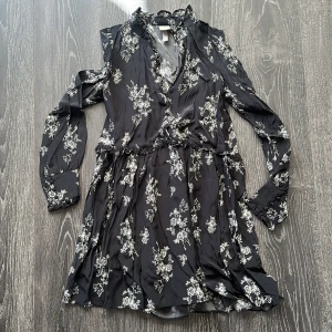 Svart blommig kortklänning från H&M - Supersöt svart kortklänning från H&M med vitt blommigt mönster. Klänningen har långärmade ärmar med knappar vid mudden, volangdetaljer vid halsen och midjan samt v-ringad omlottfront. Perfekt för dig som gillar romantiska vibes och snygga detaljer.