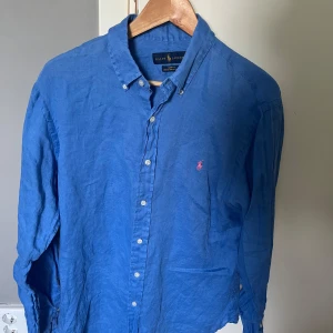 Blå linneskjorta från Ralph Lauren - Snygg blå skjorta i linne från Ralph Lauren med klassisk button down-krage och vita knappar. Långärmad modell med rosa broderad logga på bröstet. Perfekt för dig som gillar en stilren och avslappnad look.