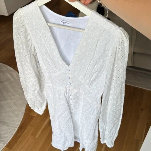 Vit kort klänning från Bubbleroom - Superfin vit klänning från Bubbleroom med broderade blommor, v-ringning och knappar framtill. Klänningen har långärmade puffärmar och är tillverkad i bomull. Perfekt för sommarens alla tillfällen, med romantisk och fräsch vibe.