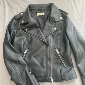 Svart skinnjacka från Glamorous - Snygg svart skinnjacka från Glamorous med klassisk bikerstil. Jackan har silvriga dragkedjor, bred krage med tryckknappar och coola detaljer på axlarna. Ingår även ett tjock skinnbälte som kom med jackan. Den är helt oanvänd.