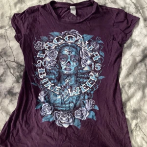 Lila Laconia Bike Week t-shirt M - Cool lila t-shirt med tryck från Laconia Bike Week 2015. Framsidan har ett stort motiv med blå och vita rosor och text, baksidan har ett mindre tryck med samma tema. Rund hals och korta ärmar. Materialet är mjuk bomull.