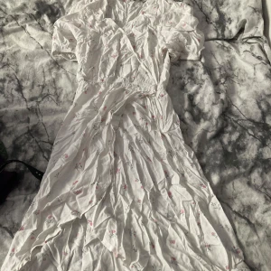 Vit blommig långklänning från 157 - Säljer en söt vit långklänning från 157 med små rosa blommor och gröna blad. Klänningen har korta ärmar, omlott-vringning och resår i midjan för en skön passform. Perfekt för varma dagar och har en romantisk vibe.