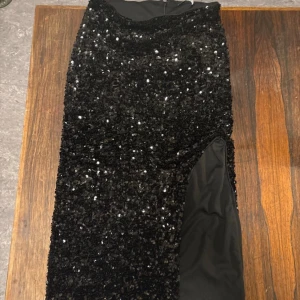 Svart paljettkjol med slits Gina Tricot - Säljer en lång svart kjol från Gina Tricot, täckt med glittriga paljetter och med en snygg slits på sidan. Kjolen har en figurnära passform och är perfekt för att sticka ut. Materialet är polyester och kjolen har dragkedja i sidan.