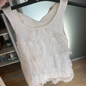 Vit topp  - Söt vit linnetopp från H&M med broderade blommor och volanger framtill. Toppen är ärmlös och har en rund halsringning. Man knäpper i halsen där bak med knappen