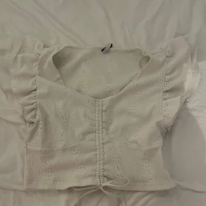 Vit blus med volangärmar från FB Sister - Supersöt vit blus från FB Sister med broderade vågiga hålmönster, volangiga korta ärmar och snörning framtill. Croppad modell med lätt struktur i tyget, perfekt för en fräsch och somrig look.