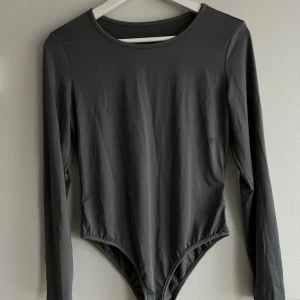Mörkgrå långärmad bodysuit - En stilren mörkgrå bodysuit med långa ärmar och rund halsringning. Tillverkad i ett stretchigt och mjukt material som sitter tight mot kroppen. Praktisk knäppning i grenen för enkel användning. Perfekt att styla med jeans eller kjol för en clean look.