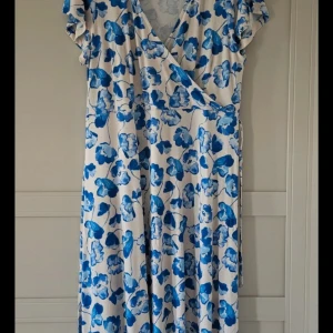 Blommig omlottklänning blå/vit L - Snygg långklänning med blått blommönster på vit botten. Omlottmodell med korta ärmar och v-ringning. Klänningen har ett flowy fall och passar perfekt för sommarens alla tillfällen. Superfräsch och lätt att styla med sneakers eller sandaler.