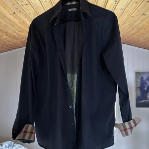 Blå Burberry skjorta med rutig detalj - Snygg blå skjorta från Burberry med klassiska rutiga detaljer på insidan av manschetterna. Skjortan har lång ärm, knappar framtill och en stilren krage. Perfekt för dig som gillar ikoniska designer. Köpt på vestaire