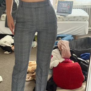 Grå rutiga kostymbyxor high waist - Snygga grå kostymbyxor med klassiskt rutigt mönster och hög midja. Byxorna har raka ben, bälteshällor och fickor fram. Perfekta för en clean och stilren look. Materialet känns mjukt och följsamt, troligtvis bomull eller polyesterblandning.