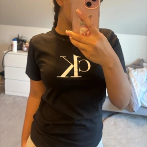 Svart CK t-shirt från Calvin Klein - Svart t-shirt från Calvin Klein med stort vitt CK-tryck på bröstet. Klassisk rund hals och korta ärmar. Mjuk bomullskvalitet som är skön mot huden. Perfekt för dig som gillar stilrena och ikoniska logotoppar.