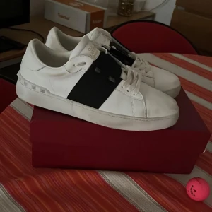 Valentino Garavani Open sneakers vit/svart - 🍀🍀🍀🍀PRUTBAR VID SNABB AFFÄR🍀🍀🍀🍀Säljer ett par snygga Valentino Garavani Open sneakers i vitt skinn med bred svart rem över mitten. Skorna har platt sula, rund tå och coola nitar på hälen. Klassisk snörning och stilren design som sticker ut. Perfekt för dig som gillar exklusiva sneakers med edge.