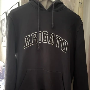 Grå hoodie från Axel Arigato - Säljer en grå hoodie från Axel Arigato med vit broderad text 'ARIGATO' på bröstet. Klassisk modell med huva, snörning och känguruficka. Perfekt för dig som gillar stilrena streetwear-plagg med en clean vibe. 100% äkta vara. Passar dig som har storlek XS-M. 