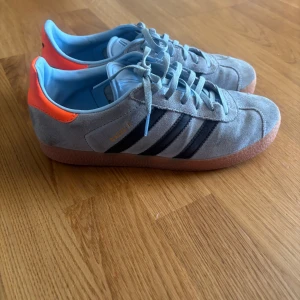 Adidas Gazelle i ljusblå mocka - Nypris 829kr.Helt slutsålda på hemsidan.Snygga Adidas Gazelle sneakers i ljusblå mocka med klassiska svarta ränder och orange detalj på hälen. Skorna har ljusblå snörning, gummisula och rund tå. Perfekta för dig som vill sticka ut med en fräsch färgkombo och ikonisk retrostil. 