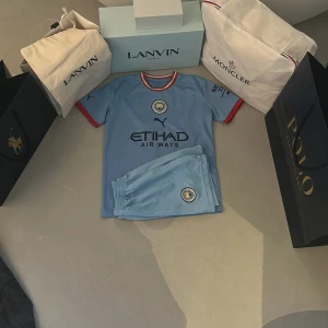 Manchester City Haaland matchställ Puma - Manchester City matchställ med Haaland 9 på ryggen. Tröjan är ljusblå med röda och vita detaljer vid kragen och ärmsluten, kortärmad och tillverkad i lätt funktionsmaterial. Setet inkluderar matchande ljusblå shorts med klubbmärke och Puma-logga.