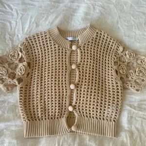 Oanvänd beige stickad kofta från Zara - Superfin beige/brunaktig stickad kofta från Zara med korta ärmar och virkade blommor på axlarna.  See-through. Koftan har genomgående knappar framtill och ett coolt nätmönster. Ribbad kant vid hals och midja. 