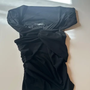 Svart omlottklänning från Bik Bok - Snygg svart kortklänning från Bik Bok i omlottmodell med offshoulder och draperade detaljer. Klänningen har en tight passform och är tillverkad i mjukt polyester-material som sitter skönt mot kroppen. Perfekt för dig som gillar stilrena och trendiga plagg.