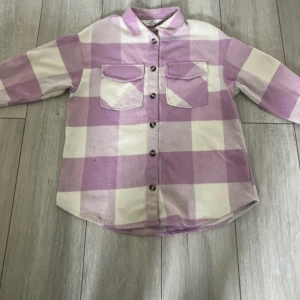 Ljuslila rutig overshirt med knappar - Overshirt i mjuk bomull med stora rutor i ljuslila och vitt. Klassisk krage, två bröstfickor med lock och knäppning framtill. Långärmad och lite oversized passform, perfekt att slänga över en t-shirt.