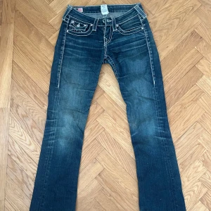 True Religion blå bootcut jeans - Snygga blå jeans från True Religion med tydliga kontrastsömmar och ikoniska bakfickor med lock och knappar. Modellen har bootcut-ben och klassisk femficksdesign. Jeansen har en cool tvätt och detaljerade sömmar som sticker ut.