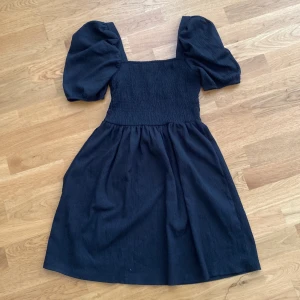 Svart puffärm klänning från H&M - Säljer en svart klänning från H&M med puffiga korta ärmar och smockad överdel. Klänningen har en fyrkantig urringning och ett mjukt, lite strukturerat tyg som ger en snygg siluett. Du står för frakten, vid mer info skriv