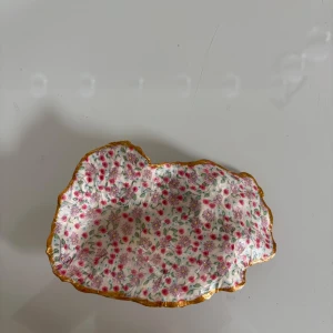 Egen smyckes skål - Bilderna visar ett dekorativt fat eller skål i form av ett snäckskal, dekorerat med blommigt mönster i rosa och lila samt guldkant. Perfekt som smyckesfat eller inredningsdetalj för dig som gillar unika och färgglada accessoarer. Passar bra i ungdomsrum eller som present. Handjort