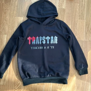 Svart Trapstar hoodie med tryck - Svart hoodie från Trapstar med stor huva och tryck i rött och grått på bröstet där det står 'Trapstar' och 'IT'S A SECRET'. Tröjan har ribbade muddar vid ärmslut och nederkant, samt en loose passform. Perfekt för dig som gillar streetwear och vill sticka ut.