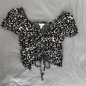 Leopardmönstrad blus från H&M - Supersöt blus från H&M med svart och beige leopardmönster. Modellen är croppad med korta puffärmar, v-ringning och snörning framtill. Smockad rygg för extra bekväm passform. Perfekt för dig som vill sticka ut med ett trendigt djurmönster.