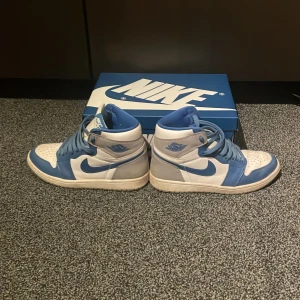  Air Jordan 1 Retro High blå/vit/grå - Säljer ett par Nike Air Jordan 1 High i blått, vitt och grått. Klassisk high-top siluett med blå swoosh, snörning och perforerad tå. Skorna är i skinn och har en ikonisk retrostil som alltid är het. Boxen medföljer och även ett par helt nya vita snören till skorna. Köpte dessa nya från Nikes SNKRS app för 2000kr men säljer dessa nu pga att de är för små.