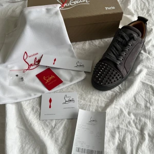 Christian Louboutin grå mocka sneakers - Säljer ett par grå sneakers från Christian Louboutin i mjuk mocka med coola svarta nitar på tån. Skorna har rund tå, platt sula och snörning. Insidan är lyxigt fodrad i beige skinn och detaljerna är riktigt snygga. Perfekt för dig som vill sticka ut med exklusiv stil.