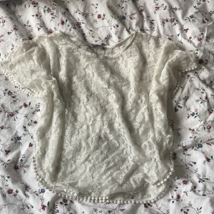 Vit spetsblus med kort ärm - Söt vit blus i spets med korta ärmar och rund hals. Blusen har fina spetsdetaljer över hela tyget och små bollar längs ärmslut och nederkant. Perfekt för en romantisk look och passar till både jeans och kjol.