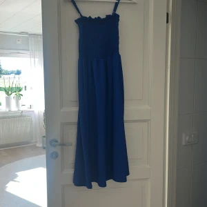 Blå långklänning med smock från H&M - Säljer en blå långklänning från H&M i storlek XS. Klänningen har smockad överdel med volangkant upptill och justerbara smala axelband. Kjolen är luftig och faller snyggt. Perfekt för dig som gillar enkel men stilren design.