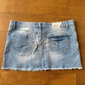 Jeans minikjol med fjärilsbroderi - Säljer en ljusblå jeans minikjol med rå kant och snyggt broderad fjäril både fram och bak. Kjolen har klassiska fickor, bälteshällor och knappstängning. Perfekt för dig som gillar Y2K-stilen och vill sticka ut med coola detaljer.