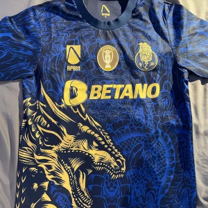FC Porto blå fotbollströja drakmotiv - FC Porto fotbollströja i mörkblått med gult drakmotiv och tryck. Tröjan har korta ärmar, rund hals och är tillverkad i lätt, ventilerande material. Framsidan pryds av klubbmärke, sponsorloggor och en stor drake. Baksidan har 'Super Bock' i gult. Storlek S men passar på M bärare också. Tröjan är använd 2-3 gånger, inte mer än så.