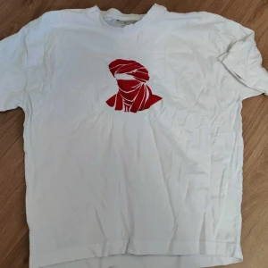 Vit t-shirt med röd tryck - Vit t-shirt med ett coolt rött tryck på bröstet som föreställer en person med turban. Klassisk rund hals och korta ärmar, perfekt för en chill och avslappnad stil. Materialet känns som bomull och t-shirten har en enkel, rak passform.