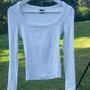 Vit långärmad topp Gina Tricot - En basic vit långärmad topp från Gina Tricot med rund, bred urringning. Toppen är ribbad och har en slim passform som sitter snyggt på kroppen. Perfekt att styla med jeans eller kjol för en clean look.