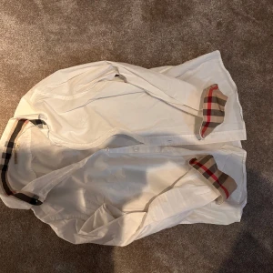 Vit Burberry skjorta med rutig detalj - Snygg vit skjorta från Burberry med klassiska rutiga detaljer i beige, svart och rött på insidan av krage och manschetter. Skjortan har knappar framtill och långärmad design. Perfekt för dig som gillar stilrena plagg med lyxig touch.