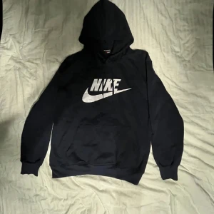 Svart Nike hoodie med stor logga - Svart hoodie från Nike med stor vit logga och swoosh på både fram- och baksidan. Tröjan har huva, magficka och ribbade muddar vid ärmslut och nederkant. Perfekt för dig som gillar sportig och avslappnad stil. Materialet är mjuk bomullsmix.