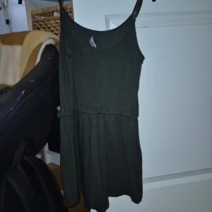 Mörkgrön kort klänning från H&M Divided - Superskön mörkgrön klänning från H&M Divided i storlek XS. Modellen är kort med smala axelband och har en dragsko i midjan för extra snygg passform. Tillverkad i mjuk bomull, perfekt för varma dagar. Enkel och stilren design som är lätt att matcha. Har byxor under