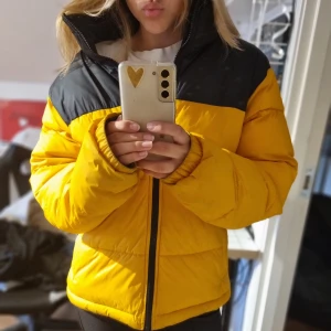 Gul och svart pufferjacka från Everest - Säljer en riktigt snygg gul och svart pufferjacka från Everest. Jackan har hög krage, dragkedja framtill och är quiltad för extra värme. Använd en vinter och mycket gott skick. Har en "reflexrem" i varje ficka.