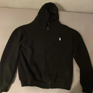 Svart hoodie från Polo Ralph Lauren - Svart hoodie från Polo Ralph Lauren med vit broderad logga på bröstet. Tröjan har dragkedja framtill, huva och är tillverkad i mjuk bomull. Custom fit-modell som sitter snyggt och passar perfekt till en avslappnad stil.