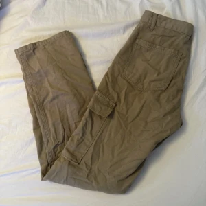 Beiga cargopants med fickor - Säljer ett par beiga cargopants med klassiska sidofickor och avslappnad passform. Byxorna har bälteshällor, knapp och dragkedja framtill samt tillverkade i mjuk bomull. Perfekta för en chill och streetig stil.