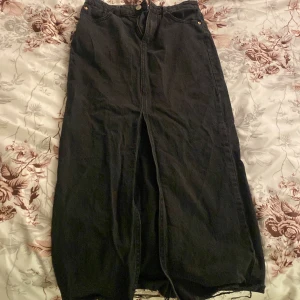 Svart jeanskjol från Stradivarius - Svart lång jeanskjol från Stradivarius i klassisk denim med hög midja, slits framtill och rå kant nertill. Kjolen har fickor både fram och bak samt dragkedja och knappstängning. Perfekt för en cool och avslappnad look.