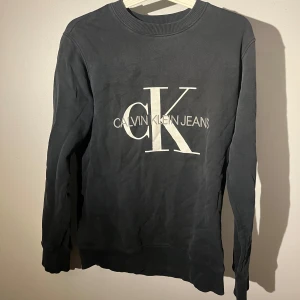 Svart sweatshirt från Calvin Klein Jeans - Svart sweatshirt med stor vit CK-logga och Calvin Klein Jeans-tryck på bröstet. Tröjan har rund hals, ribbade muddar och är tillverkad i mjuk bomull. Perfekt för dig som gillar stilrena och klassiska märkesplagg. Sitter mer som xss