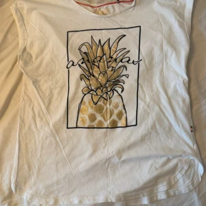 Vit t-shirt med ananastryck - Vit t-shirt med coolt tryck av en ananas i guld och svart, inramad med svart linje och text över motivet. Modellen är ärmlös och har rund hals. Materialet känns som mjuk bomull, perfekt för chill dagar. Snygg och unik design som sticker ut.
